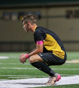 Soccer player in a black and yellow jersey crouching on field, wearing a pink captain's armband during a match.