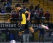 Soccer player in black and yellow uniform dribbles ball during night match on field with audience in background.