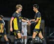 Two soccer players in black and yellow jerseys shaking hands on the field during a night game.