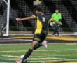 Soccer player in yellow and black uniform taking a shot at goal during a night match.