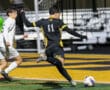 Soccer player in black and yellow jersey kicks ball towards goal on a colorful field during a match.