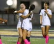 Soccer players celebrating a goal, wearing white uniforms and pink socks on the field at night.