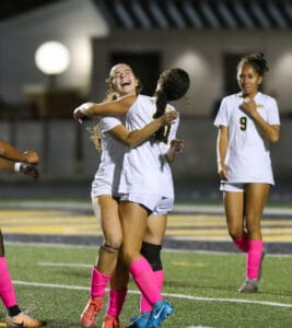 Soccer players celebrating a goal, wearing white uniforms and pink socks on the field at night.