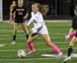 Soccer player in white uniform dribbles the ball on a field, focused on the game under bright stadium lights.
