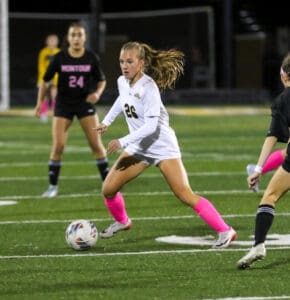 Soccer player in white uniform dribbles the ball on a field, focused on the game under bright stadium lights.