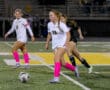 Female soccer players in white jerseys and pink socks playing on a field during a night match.