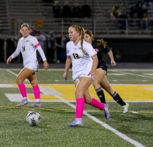 Female soccer players in white jerseys and pink socks playing on a field during a night match.