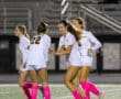 Four female soccer players in white jerseys celebrating on the field at night.