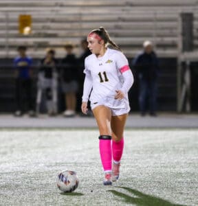 Soccer player in white kit and pink socks and headband with ball on the field at night game.