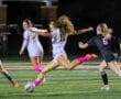 Soccer player in white kicks ball during nighttime match against opposing team in black.