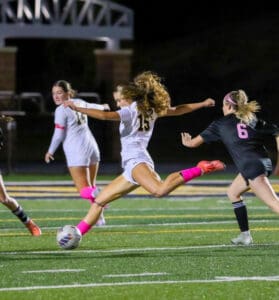 Soccer player in white kicks ball during nighttime match against opposing team in black.