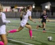 Soccer player in white uniform dribbles ball, pursued by opponent in black, under stadium lights.