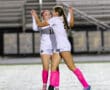 Two female soccer players in white jerseys and pink socks celebrating on the field at night.