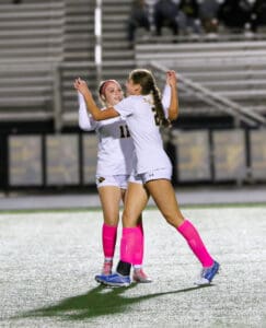 Two female soccer players in white jerseys and pink socks celebrating on the field at night.