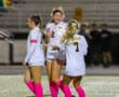 Three female soccer players in pink socks celebrate on the field during a night game.