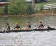 Rowing team of five in black gear on a river, passing by greenery and moored boats.