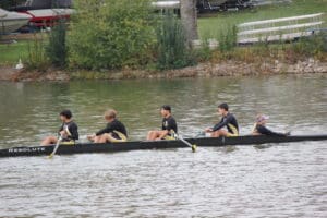Rowing team of five in black gear on a river, passing by greenery and moored boats.