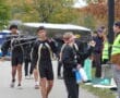 Rowers in black uniforms carrying a boat on a leafy pathway during autumn.