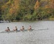 Four-rower team paddling on a scenic lake surrounded by autumn trees, showcasing teamwork and nature's beauty.