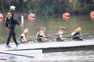 Rowers prepare their boat on a calm lake, guided by a coach, with trees and safety buoys in the background.