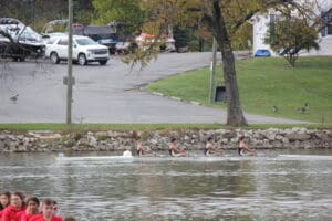 Rowing team competes on a calm lake with cars and geese visible on the shore in the background.