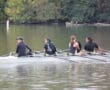 Rowing team practices on calm river, surrounded by lush trees, under a clear sky.