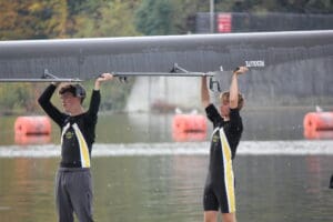 Rowing team members carrying a wet boat on the water's edge, wearing black uniforms with yellow and white stripes.
