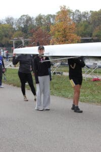 Rowing team carrying boat on riverside path, autumn trees in background.