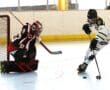 Roller hockey player in white attempts shot against goalie in black during an indoor game.