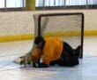 Hockey goalie in orange jersey makes a save on the ice rink, lying in front of the net.