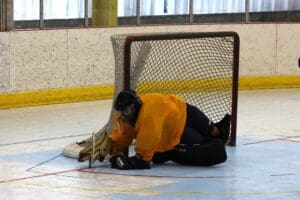 Hockey goalie in orange jersey makes a save on the ice rink, lying in front of the net.