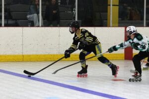 Roller hockey players in action, one in black and yellow, skating fast on rink, with spectators in background.