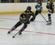 Roller hockey game action with players in black and green jerseys competing for the puck on an indoor rink.