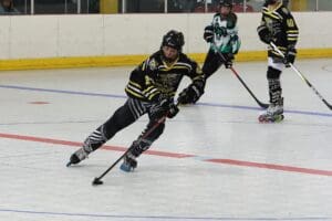 Roller hockey game action with players in black and green jerseys competing for the puck on an indoor rink.
