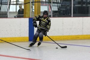 Inline hockey player in black and yellow gear skates on a rink, controlling the puck with a stick.