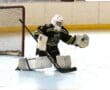 Hockey goalie in action blocking a puck on an indoor ice rink with goal net in the background.