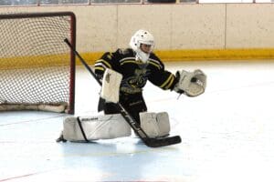 Hockey goalie in action blocking a puck on an indoor ice rink with goal net in the background.