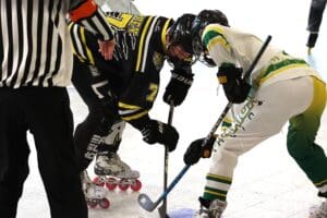 Roller hockey players face-off as referee watches, wearing team jerseys and gear on the rink.