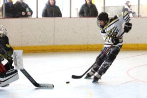 Hockey player in action on the rink with goalie ready, spectators watching from behind the barrier.