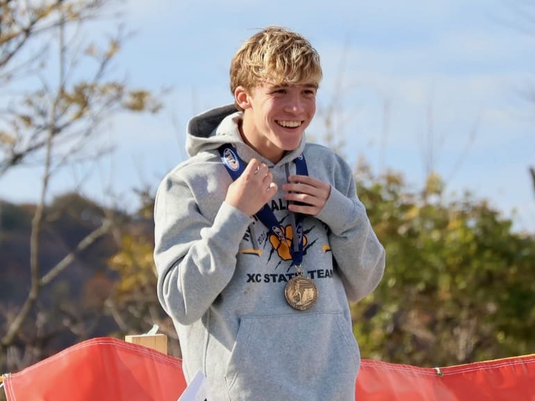 Young athlete proudly wears a medal while smiling at an outdoor award ceremony, wearing a gray hoodie.