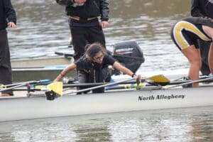 Team at rowing competition aligning oars on water, North Allegheny boat in focus, sunny outdoor setting.