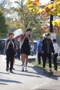 Rowing team carrying a boat at a regatta, surrounded by autumn trees and spectators.