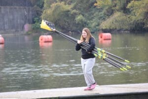 Woman carrying rowing oars on dock by the lake with orange buoys in the background.