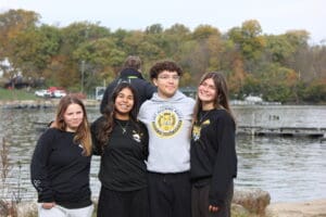 Group of smiling friends by a lake in autumn, wearing casual outfits with trees in the background.