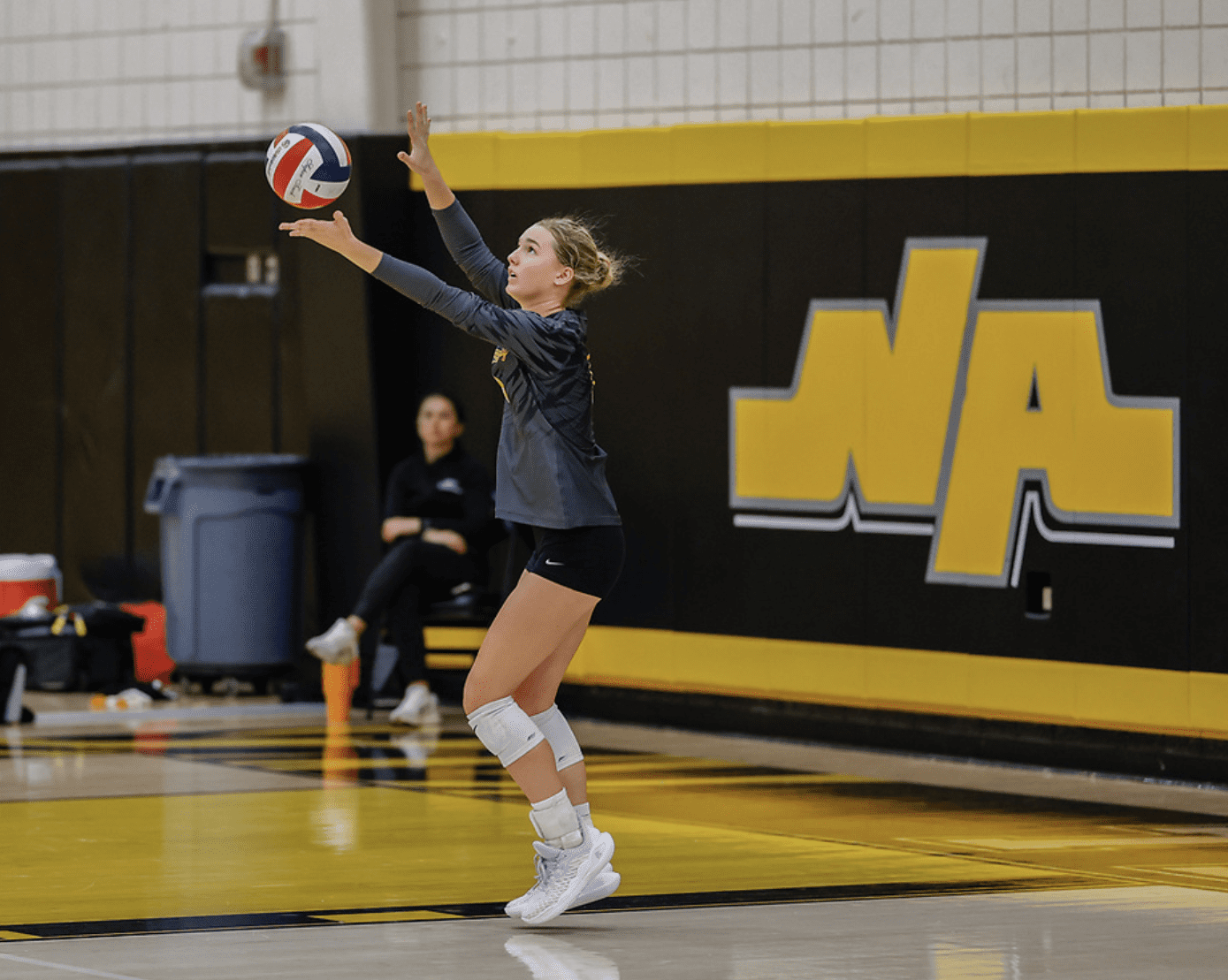 Volleyball player jumps to serve at indoor court during a game, wearing knee pads and athletic shoes.