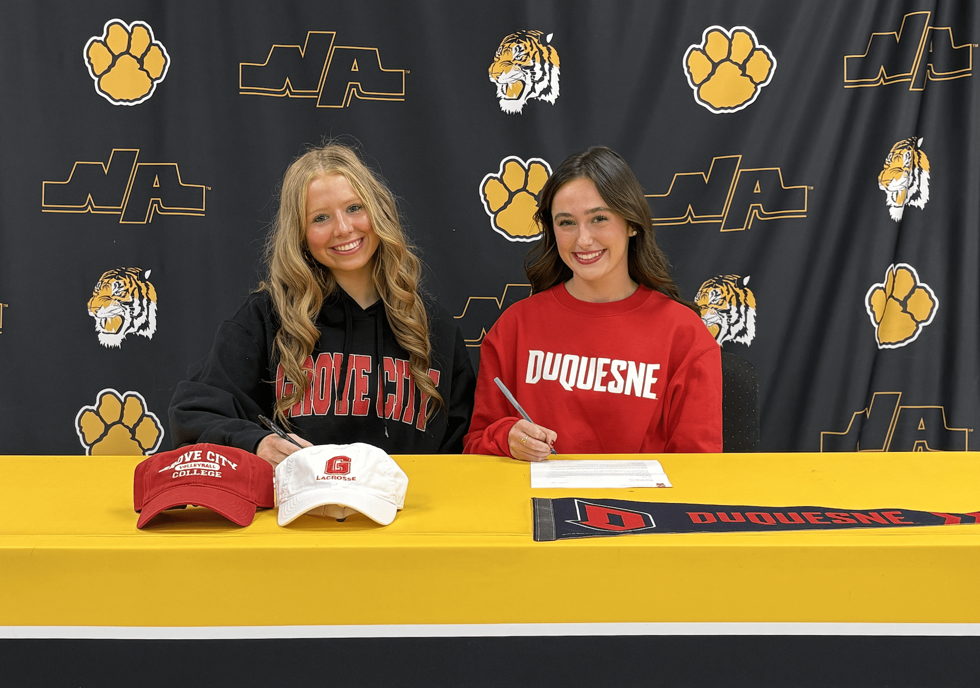 Two students in collegiate sweatshirts signing documents at a table with Grove City and Duquesne logos.
