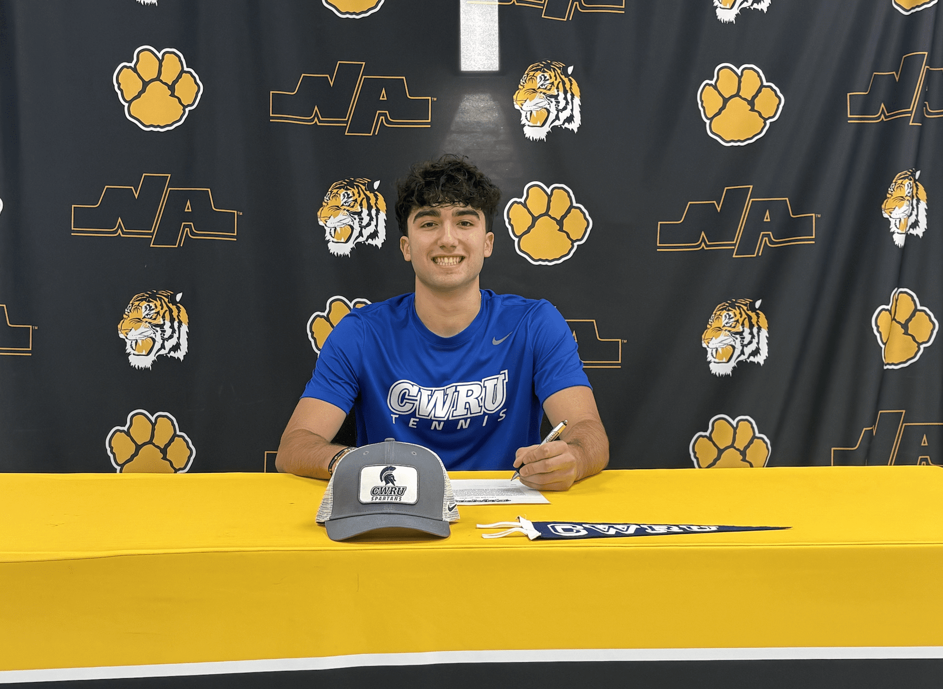Young athlete signs with CWRU Tennis, smiling at a desk with sports backdrop and cap.