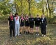 Rowing team celebrates outdoors with medals, smiling under trees, dressed in sports attire.