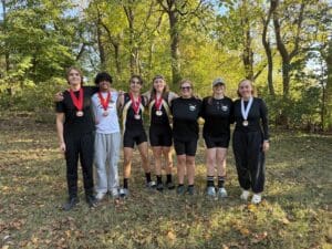 Rowing team celebrates outdoors with medals, smiling under trees, dressed in sports attire.