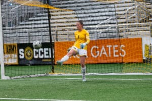 Soccer goalie in yellow jersey kicks ball during match, in front of net on green field.